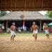 Ilustrasi tiga pemuda mengenakan pakaian tradisional dan kepala ikat sedang bertanding Sepak Takraw di lapangan beralaskan tanah dengan latar bangunan beratap rumbia serta bendera Indonesia